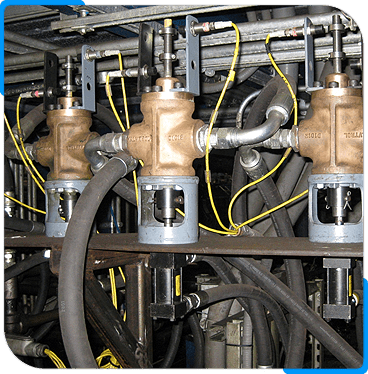 Three industrial valves with attached hoses and yellow electrical wires are mounted in a metal framework. The valves are made of brass and steel, and various pipes and cables run around and behind them.