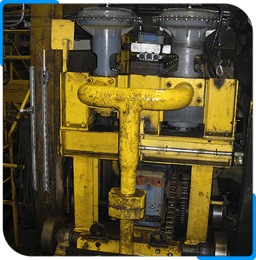 A close-up view of a heavy-duty industrial machine with yellow metal parts, valves, gears, and pipes, situated in a factory or workshop environment.