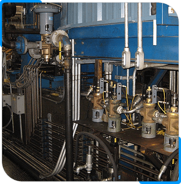 A close-up view of industrial machinery with metal pipes, valves, gauges, and wiring, all mounted on and around a blue-painted machine in a factory or plant setting.