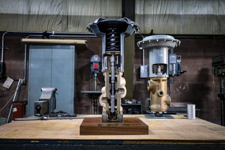 A mechanical valve assembly is displayed on a wooden table in an industrial workshop, with machinery, tools, and equipment visible in the background. The valve is cut open to reveal its internal components.