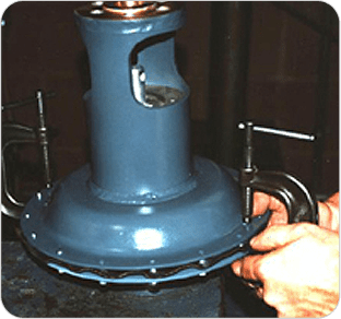 A person uses a C-clamp to secure or tighten a blue metal pump housing or casing, with bolts visible around the edge of the circular base.